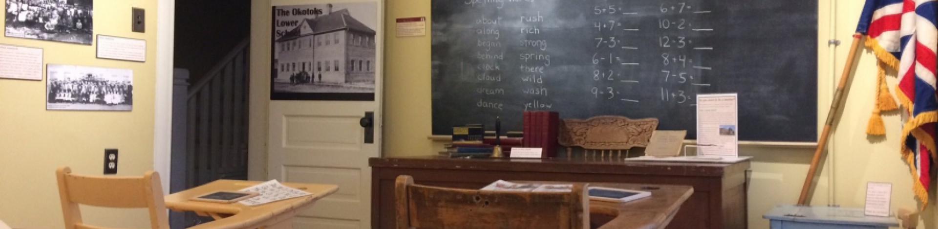 Schoolroom set up in the Okotoks Museum with blackboard, flag, and desks.