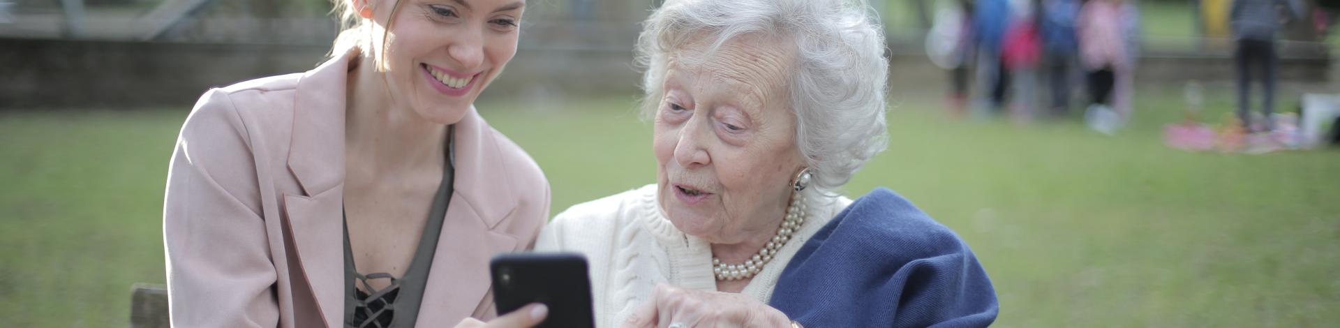 Senior woman sitting on bench in park with younger woman while looking at a phone