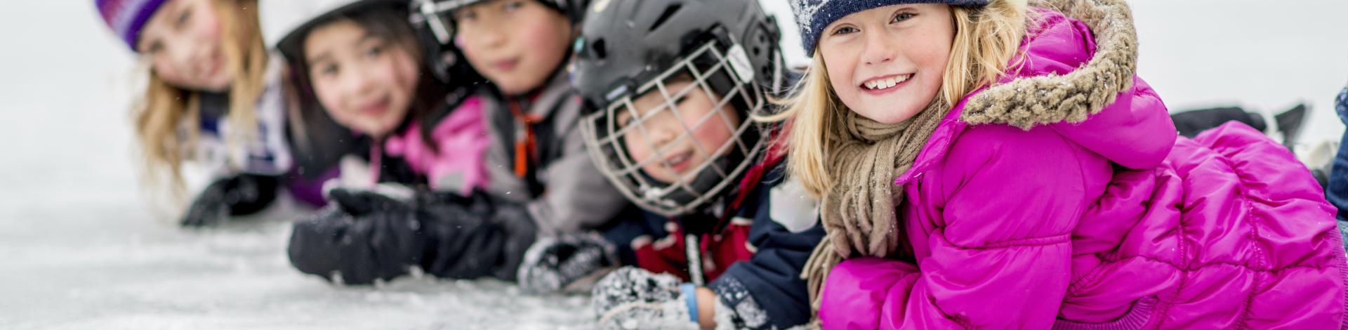 Line of children in helmets and skates laying on outdoor skating rink