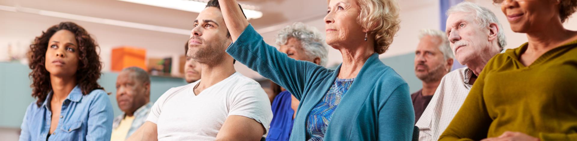 Women with blonde hair wearing a blue cardigan sits amongst other adults in a crowd with her hand up 