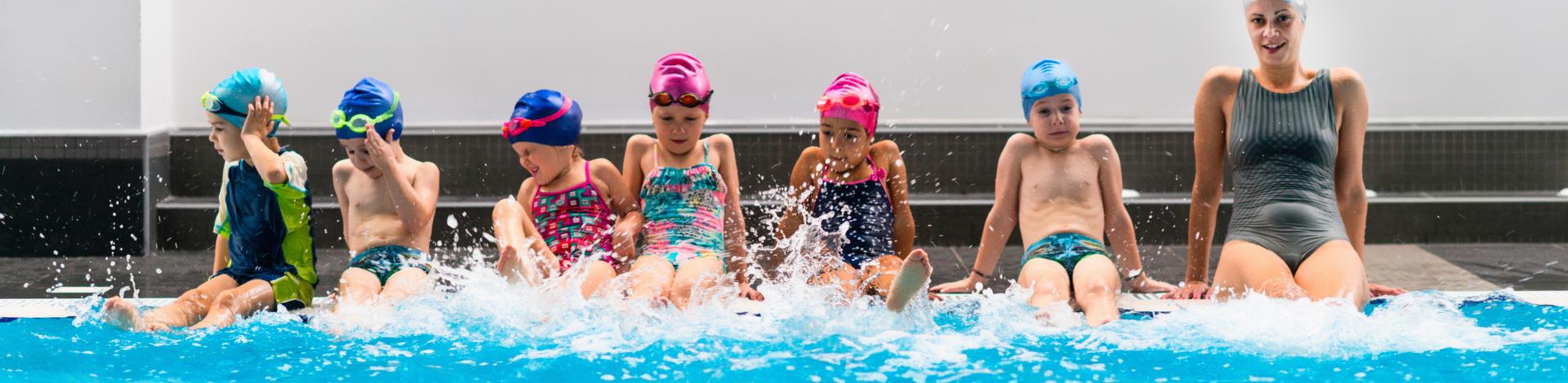 group of children participating in swim lessons splashing legs in water while sitting on pool deck