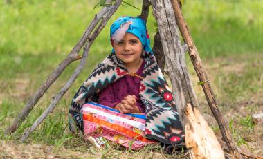 Indigenous Girl in colourful traditional dress sitting on the grass underneath poles fastened in the style of a Tipi