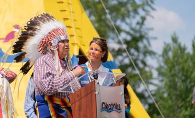 Mayor Thorn and an Elder in traditional head dress standing at a podium during a speech at the Tipi transfer ceremony. The yellow Tipi is in the background.