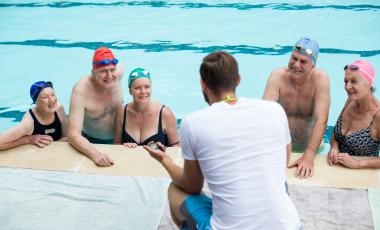 rear view of male instructor assisting senior swimmers poolside